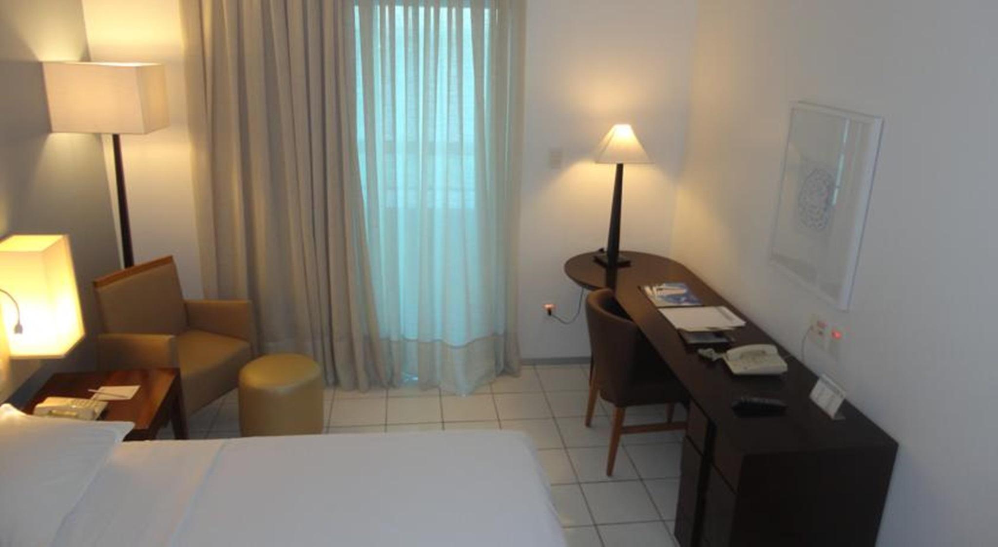 Hotel Luzeiros Fortaleza Exterior foto The photo shows a hotel room with a modern and minimalist design. It includes a bed with white linens, a small seating area with two chairs and a round ottoman, and a desk with some stationery items and a telephone. A lamp stands on the desk, providi