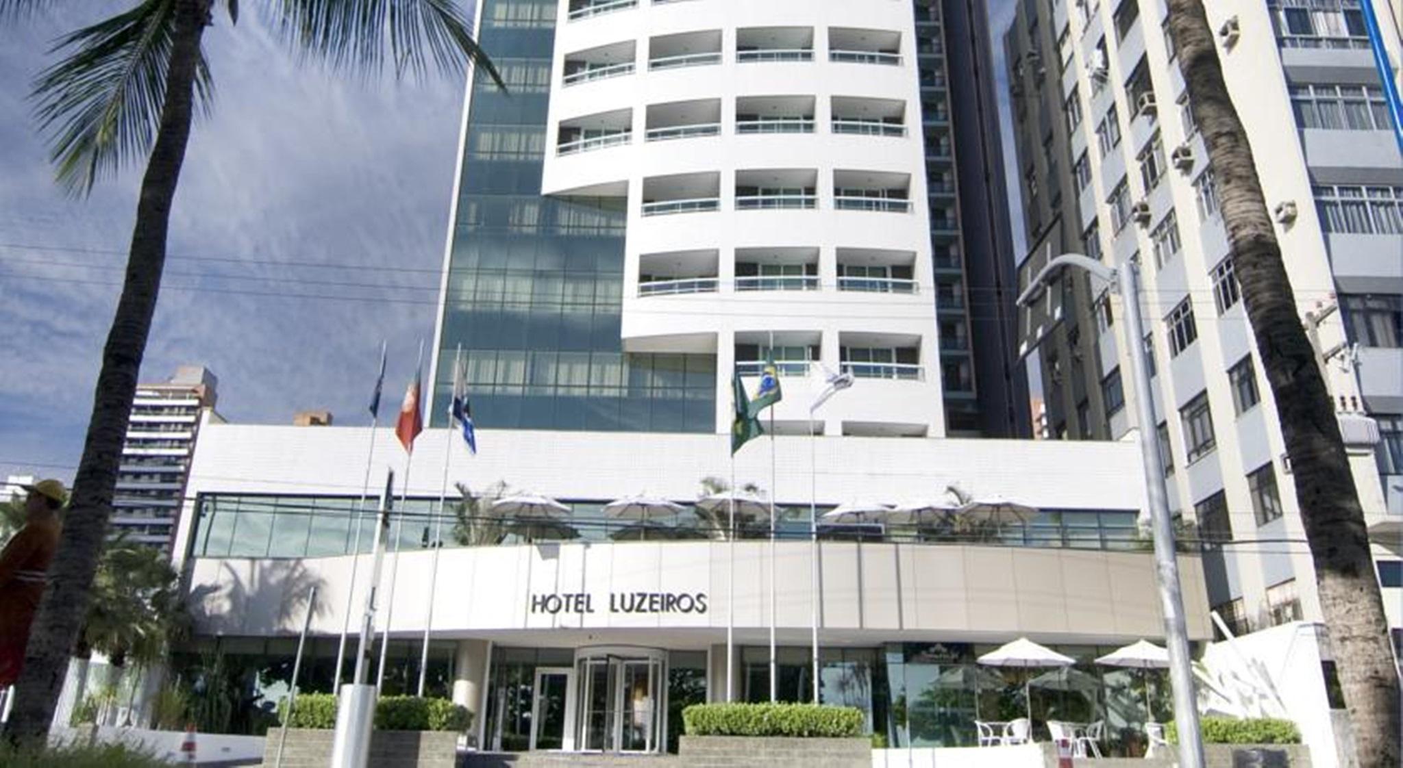 Hotel Luzeiros Fortaleza Exterior foto The photo shows the facade of a modern hotel named "Hotel Luzieros." The building features a sleek design with a combination of glass and concrete elements. Several flags are displayed in front, suggesting an international appeal. The entrance area i