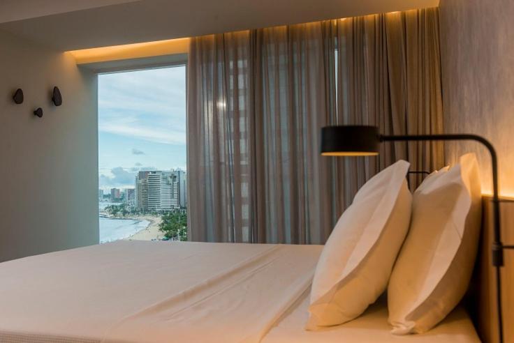 Hotel Luzeiros Fortaleza Exterior foto The photo shows a modern and stylish bedroom. The bed is adorned with two large, fluffy pillows, and the bedding appears to be neatly arranged. A warm light fixture is mounted on the wall beside the bed, adding a cozy ambiance. There are large window