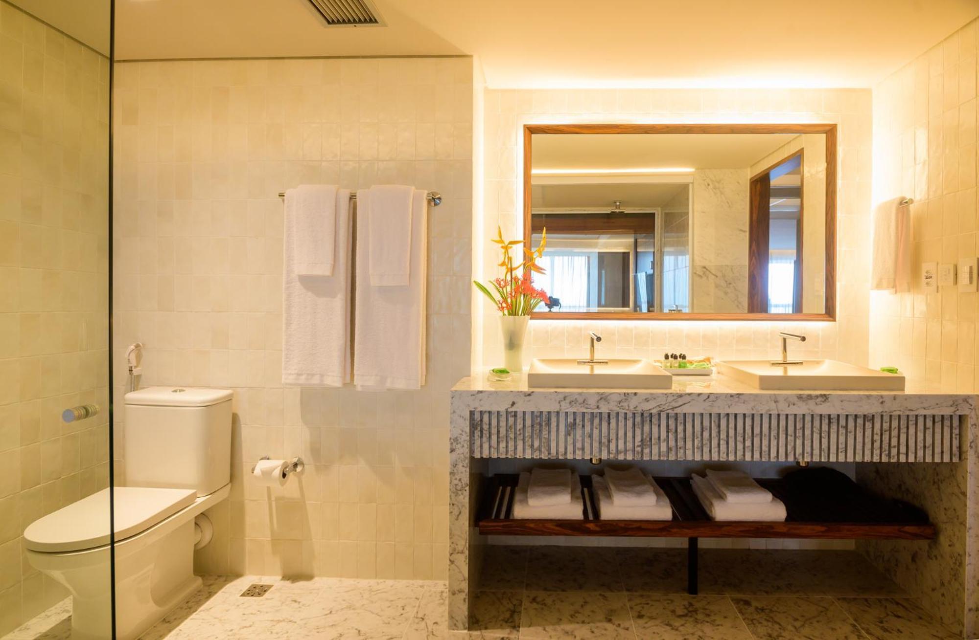 Hotel Luzeiros Fortaleza Exterior foto The photo shows a modern bathroom. It features a glass shower enclosure, a white toilet, and a double sink vanity made of stone. Above the sink, there's a large mirror with warm lighting around it. Hand towels hang neatly on a rack, and there are som