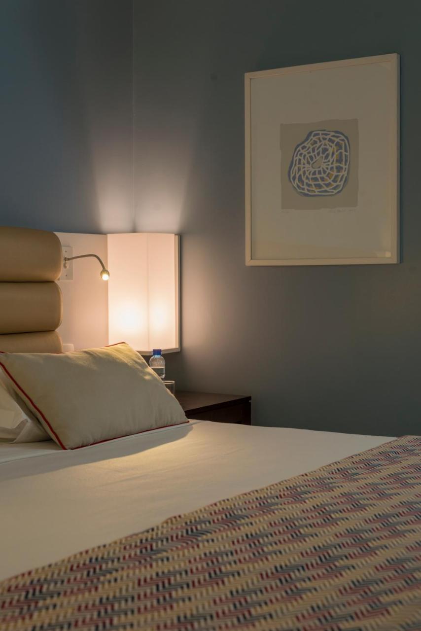 Hotel Luzeiros Fortaleza Exterior foto The photo shows a cozy bedroom setting. There is a neatly made bed with a white sheet and a couple of decorative pillows. One of the pillows is slightly displaced. On the bedside table, there is a small bottle of water and a lamp with a warm glow. Th