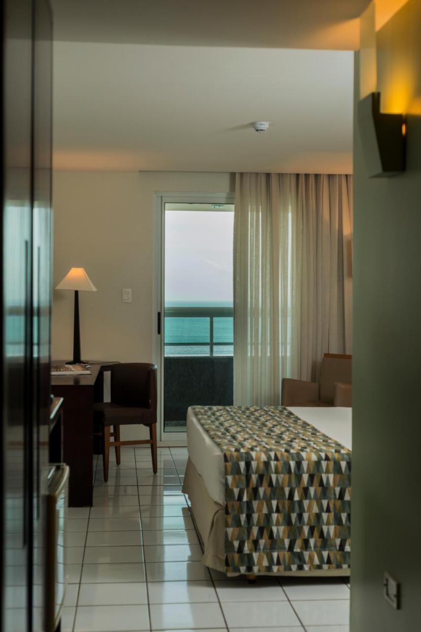 Hotel Luzeiros Fortaleza Exterior foto The photo showcases a hotel room with a balcony view. It features a bed with a patterned blanket, a bedside table, and a lamp. There is a small wooden desk with a chair in the corner. Sheer curtains frame a sliding glass door that opens to the balcon