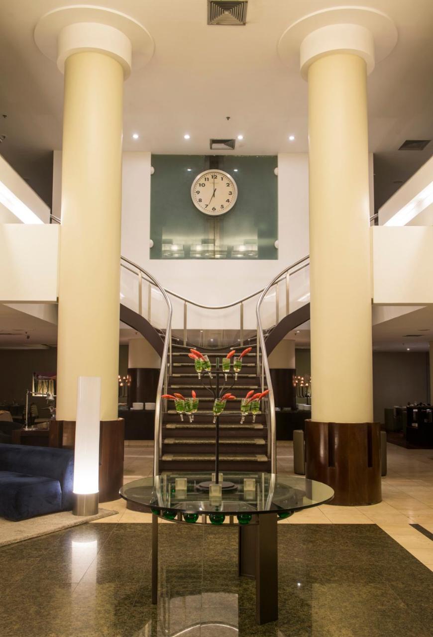 Hotel Luzeiros Fortaleza Exterior foto The photo shows an elegant interior of a hotel lobby. There is a grand, curved staircase in the center, leading to an upper level. The staircase is flanked by large columns, and there are decorative plants or floral arrangements at its base. Above th